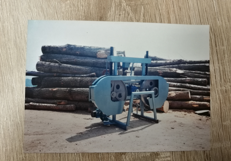 Stara tračna žaga za razrez hlodovine postavljena na prostem pred skladovnico lesenih hlodov.