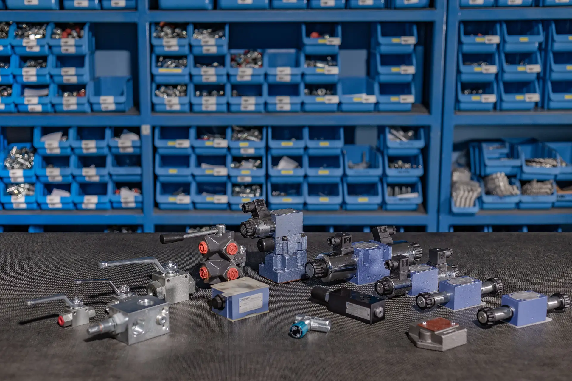 Hidravlični ventili, ročni upravljalniki in industrijski priključki razstavljeni na mizi pred skladiščnimi regali.